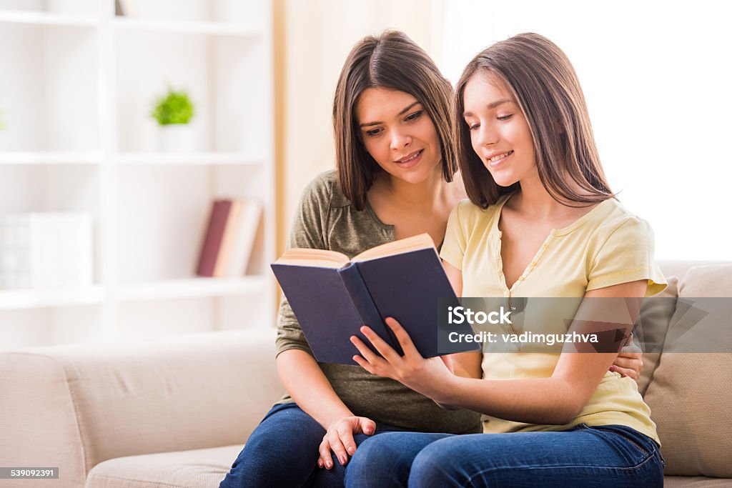 Mother and daughter Beautiful mother and her cute teen daughter are reading a book together while sitting on sofa at home. Book Stock Photo