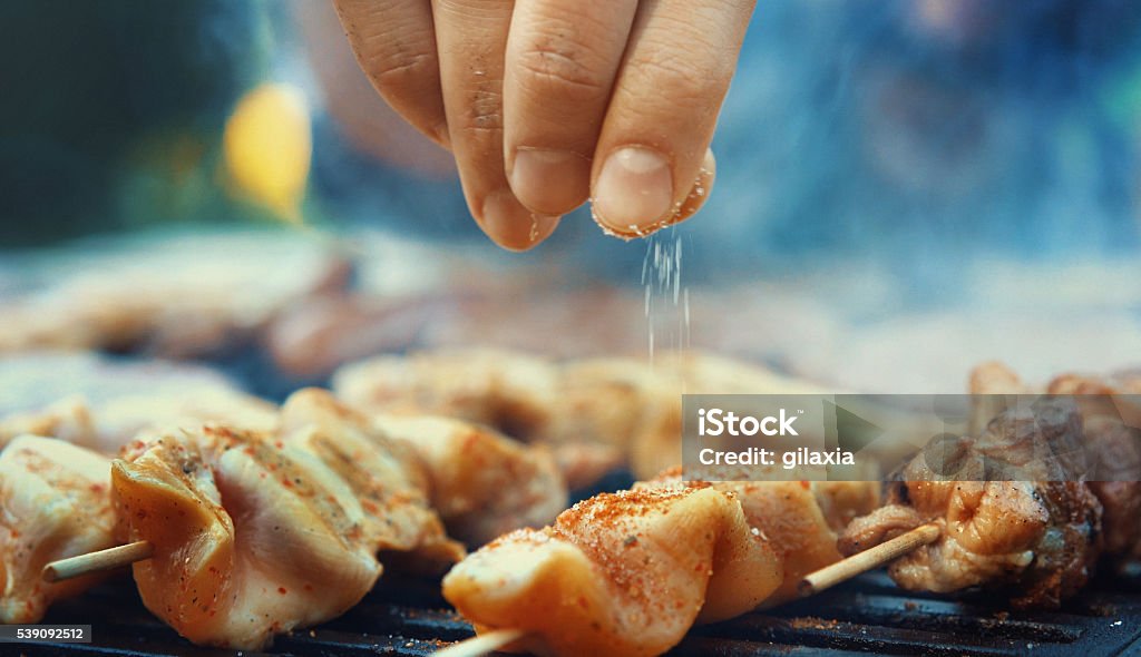 Chef seasoning meat on a grill. Closeup of unrecognizable male chef seasoning meat on a grill with a pinch of salt. Very shallow focus. Salt - Mineral Stock Photo