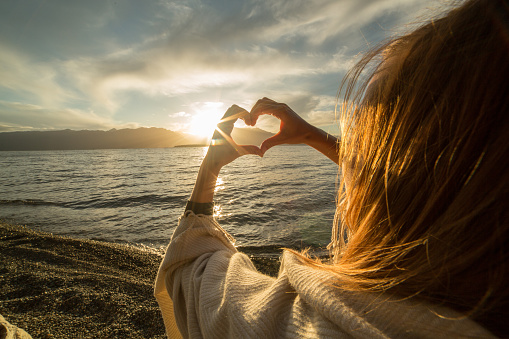 Young woman by the lake making a heart shape finger frame. Lake and mountain landscape.