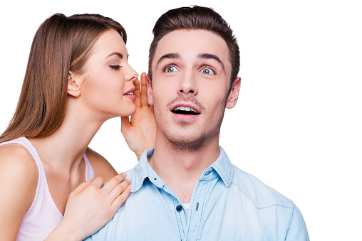 Beautiful young women holding her mouth near ear of her boyfriend while standing against white background
