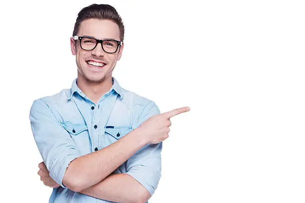Handsome young man in shirt looking at camera and pointing away while standing against white background