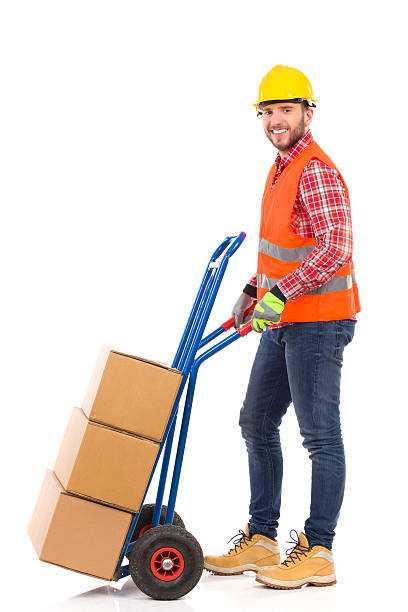 Mover with a push cart. Smiling man in yellow hardhat and orange reflective vest pushing a delivery cart. Full length studio shot isolated on white. mover delivery person box hand truck stock pictures, royalty-free photos & images
