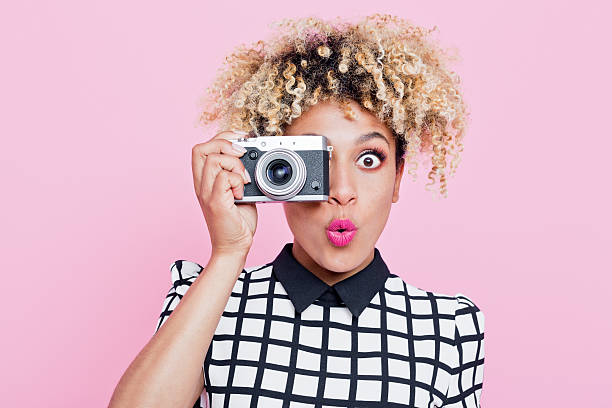 Surprised young woman wearing sunglasses, holding camera Portrait of surprised beautiful afro american young woman wearing grid check playsuit, holding a camera in hand, looking at camera. Studio shot, one person, pink background. dress photos stock pictures, royalty-free photos & images