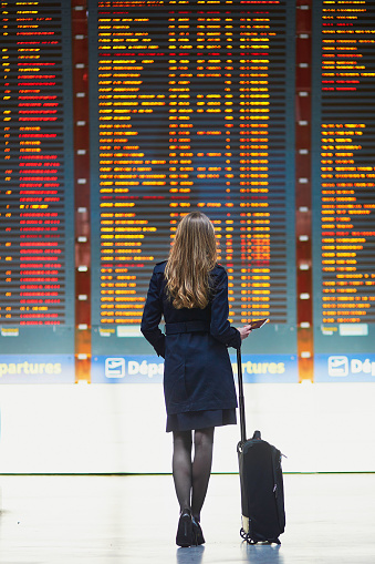 Young woman in international airport looking at the flight information board, holding passport in her hand, checking her flight