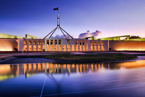 Australian national parliament house in Canberra. Facade of the buidling brightly illuminated and reflecting in blurred water of fountain pond under waving national flag on flagpole at sunset.