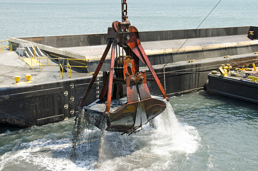 A cable powered crane uses a clam shell bucket to remove sand in a dredging operation in Port Orford Oregon