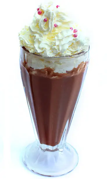 Photo showing a single chocolate milkshake, isolated against a white background.  The drink has been topped with whipped cream / spray cream and sprinkles, being served in a tall American diner-style 'knickerbocker glory' glass.