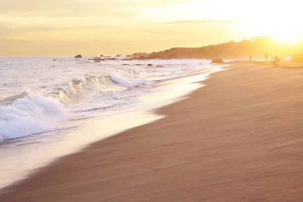 A DSLR photo of a warm orange sunset at the beach. Vanishing point of the rolling waves. The water is green with white foam and sun glare. Yellowish sand in the foreground. Side view of the shore. A silhouette of a hill with some beach houses is at the background where the sun comes down. The sun is yellow and glowing above the hill at the right corner of the photo. At the distance there are small silhouettes of fisherman fishing at the beach. The sky is cloudy with orange colours. 