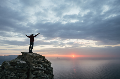 man raises his arms on top of the cliff on the sea front.