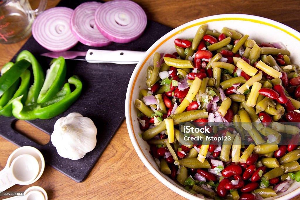 Bowl of three bean salad and ingredients. Food preparation. Bowl of three bean salad with ingredients. Green beans, kidney beans, onions, garlic, bell pepper.  Measuring spoons, knife, cutting board. High angle view. Wax Bean Stock Photo