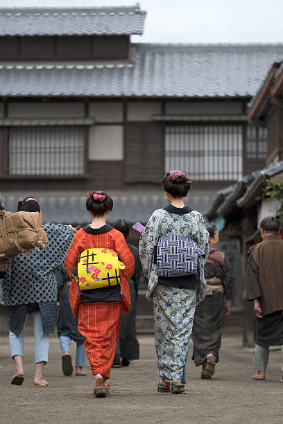 A large group of Japanese women and men walk down a traditional street.  Women are wearing kimonos and men are wearing period clothing.  Toei Kyoto Studio Park.  Kyoto, Japan.