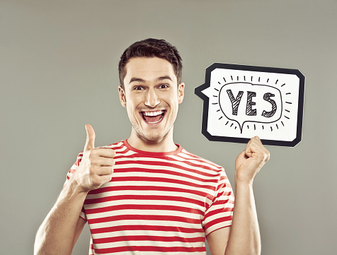 Portrait of excited young man wearing striped t-shirt, holding in hand a speech bubble with drawn word yes, staring at camera with mouth open and thumb up. Studio shot, grey background.