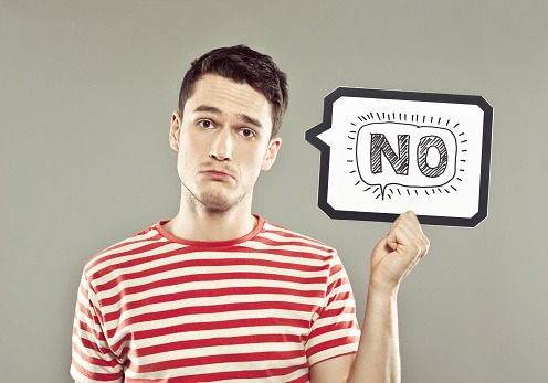 Portrait of sad young man wearing striped t-shirt, holding a speech bubble with drawn word, looking at camera. Studio shot, grey background.