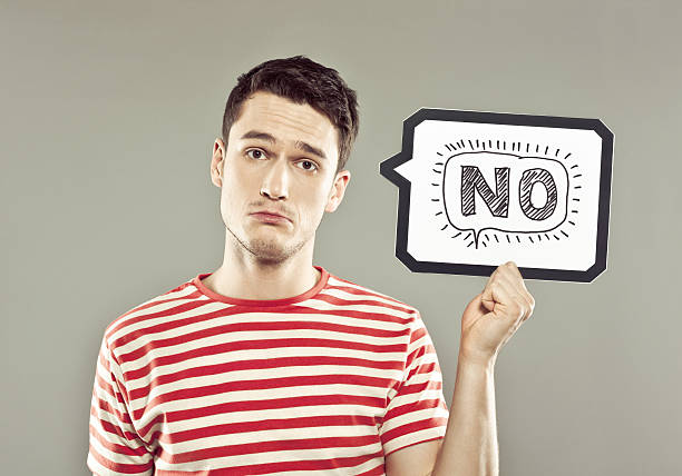 Young man holding speech bubble with word no Portrait of sad young man wearing striped t-shirt, holding a speech bubble with drawn word, looking at camera. Studio shot, grey background. single word no stock pictures, royalty-free photos & images