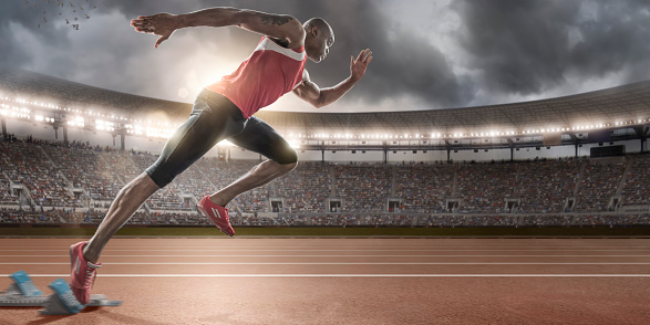 Sprinter bursting out of blocks on outdoor running track. Action set in generic outdoor floodlit athletics stadium full of spectators under dramatic stormy evening sky. Athlete is dressed in generic athletics strip and running spikes. 