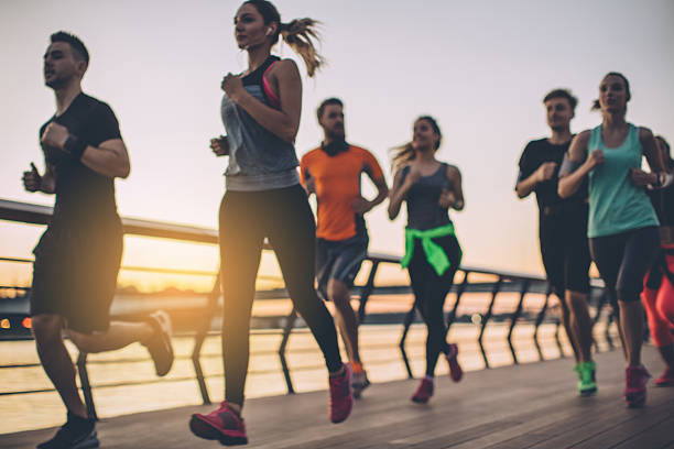 Energy is contagious. Group of young people competing in a race. Young men and women running on riverside promenade at sunset. They are wearing sport clothing. marathon stock pictures, royalty-free photos & images