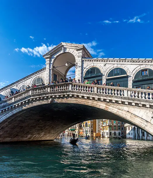 Scenic view of the centre of the Rialto Bridge on the Grand Canal, Venice, Italy with a crowd of tourists looking over the balustrades a gondola below and a glimpse of historic palazzi behind