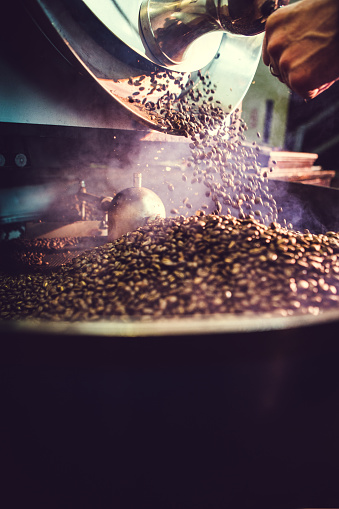 The process of roasting a batch of high quality single origin coffee beans in a large industrial roaster.  A mans hand is visible releasing the roasted beans into the cooling cycle.  Smoke rises from the hot beans.  Vertical with slight motion blur and copy space.  