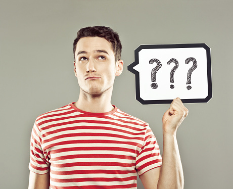 Portrait of young man wearing striped t-shirt, holding a speech bubble with drawn question marks, looking up. Studio shot, grey background.