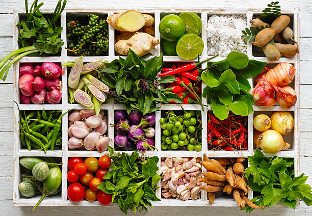 Thai food herbs and spice ingredients in a wooden tray. Main market fresh Thai food herbs and spice ingredients in a colorful arrangement in an old vintage wooden compartment tray on an old wooden table. Ingredients include lemongrass, kaffir lime leaves, galangal, finger root, dried and fresh chili peppers, Thai aubergines, garlic, coriander, holy basil, sweet basil, tamarind, peppercorns, spring onion, onion, tomato, lime and coconut. organic spice stock pictures, royalty-free photos & images