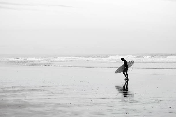 Surfer Exiting the Ocean An image of the silhouette of a surfer leaving the ocean on a winter morning in Pismo Beach, California. black and white beach stock pictures, royalty-free photos & images