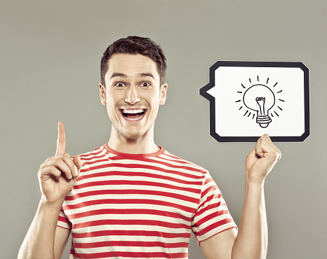 Portrait of excited young man wearing striped t-shirt, holding  in hand a speech bubble with drawn light bulb, staring at camera with mouth open. Studio shot, grey background.