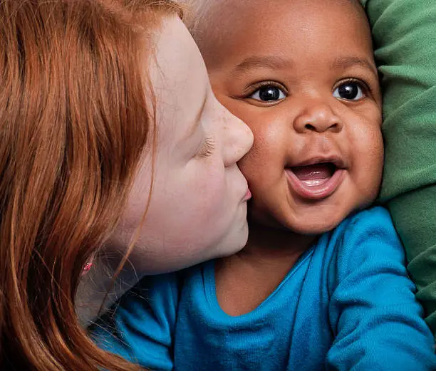 Photo of little redhead girl kissing smiling adopted african ethicity baby sister
