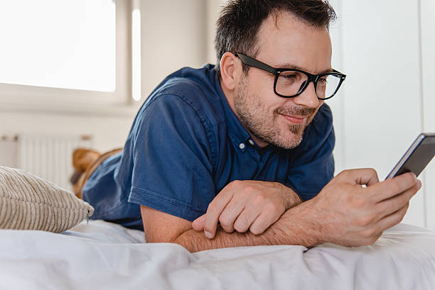 homem usando telefone inteligente na cama - mid adult men smart casual white happiness - fotografias e filmes do acervo