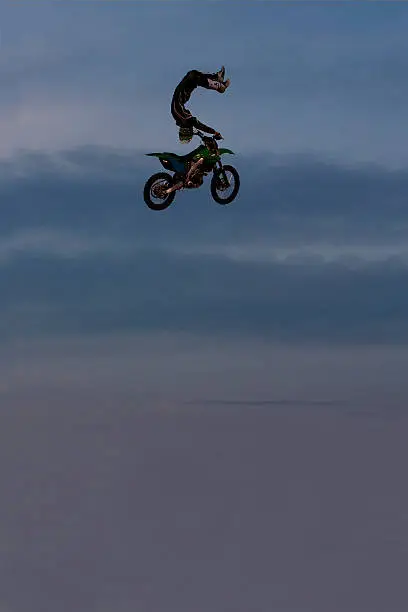A male freestyle FMX motocross rider does a trick off a big jump at night. 