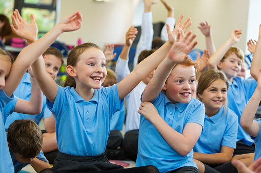 Excited school children in school uniform with hands up ready to answer a question from the teacher