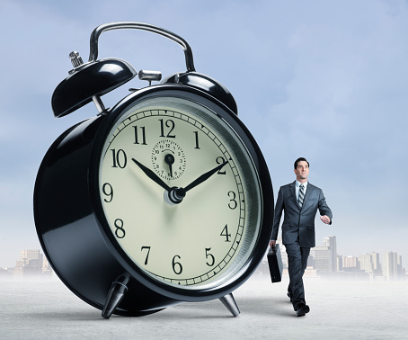 A businessman walks away from a big city skyline in the distance and past a very large alarm clock. He is carrying a briefcase and is very determined in his walk.  He is obviously on a deadline. The time on the clock is 10:10 am.