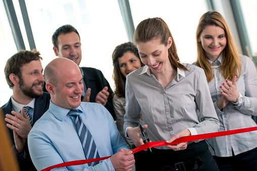 Businesspeople in the office. Woman is cutting a red ribbon with scissors, man is helphing her to hold it, other people are applauding. They are all happy with smile on their faces.