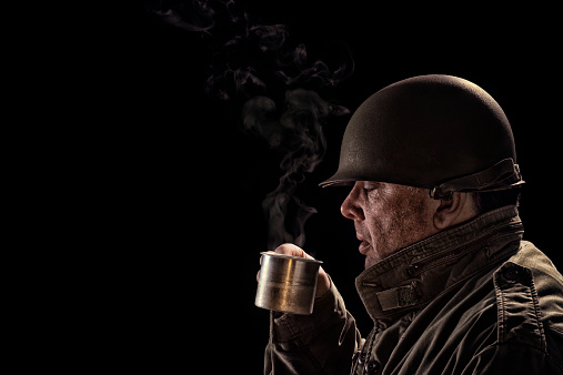 Vintage Army Soldier drinking hot coffee from a tin cup against a black background, with low key lighting