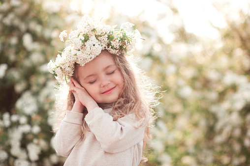 Smiling baby girl 3-4 year old posing in meadow over flowers. Wearing floral wreath outdoors. Childhood.
