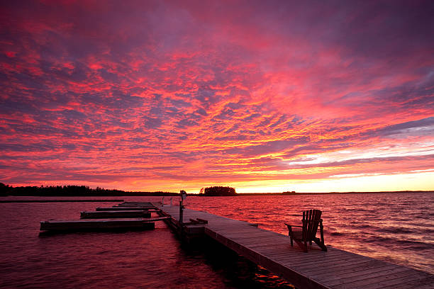 An adirondack chair on a dock by a lake with a gorgeous sunset. The location is Muskoka region in Ontario, Canada. Summer is prime time in cottage country and this region is a prime destination for Toronto residents who want to get away from the city. The dramatic sunset is not digitally enhanced. It was one of the prettiest sunsets this photographer has seen.