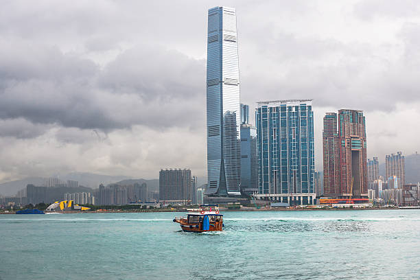 A traditional junk boat navigates the waters of Victoria Harbour, surrounded by Hong Kong's impressive skyscrapers. The scene captures the blend of culture and modernity.