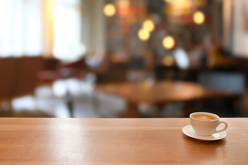 Cup of coffe on a wooden table in a cafeteria or restaurant. Plenty of copy space in the middle of the empty table. Bright light coming from the windows, with defocused background. Citylife and modern lifestyle.