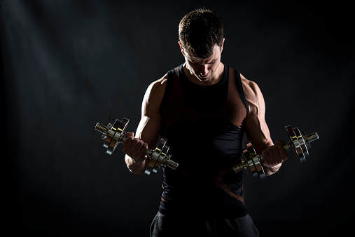 Muscular young man doing exercises for biceps muscles with dumbbells on black background. Looking down.