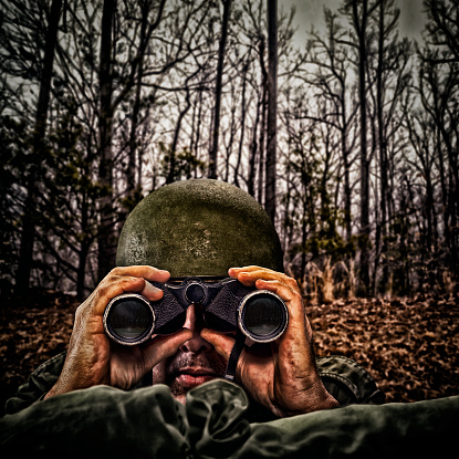 Vintage Army Soldier looking through binoculars behind sand bags of his fox hole at the edge of the forest
