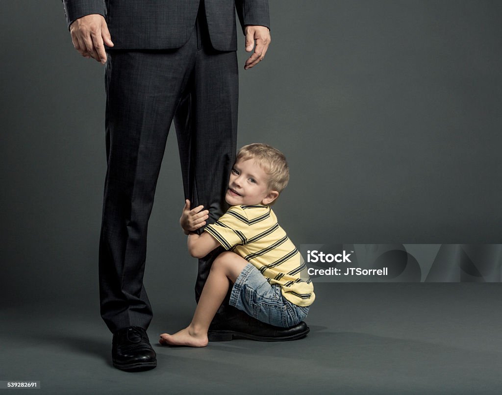 Fatherhood and Work Balance An adorable young boy clings to his father's leg who is dressed in a suit for work. 2015 Stock Photo