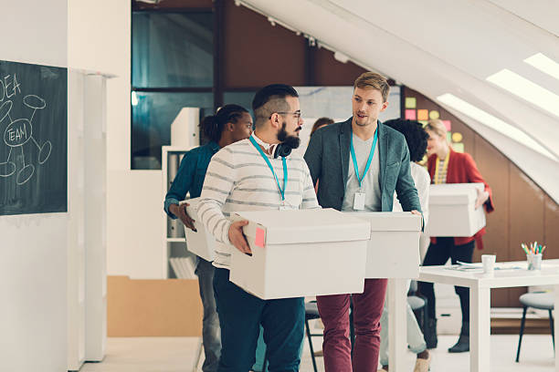 Startup Business Team Moving To New Office. Group of young creative people moving to their new startup business office. They all carry boxes as a team. Selective focus to two men in foreground talking while carry boxes. company relocation stock pictures, royalty-free photos & images