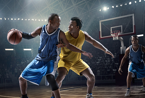 Low angle view of a professional basketball game. A player is trying to block  the opposite team player with a ball. A game is in a indoor floodlit basketball arena. All players are wearing generic unbranded basketball uniform.