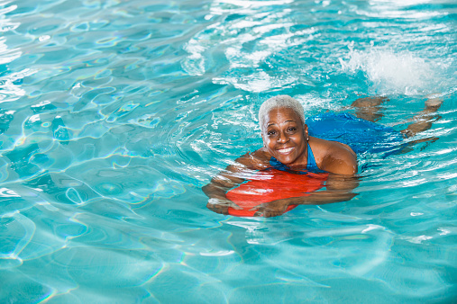 High angle view of a senior African American woman (in her 60s) swimming in a pool hanging onto a red kickboard as she kicks her legs.   She is having fun, smiling at the camera.