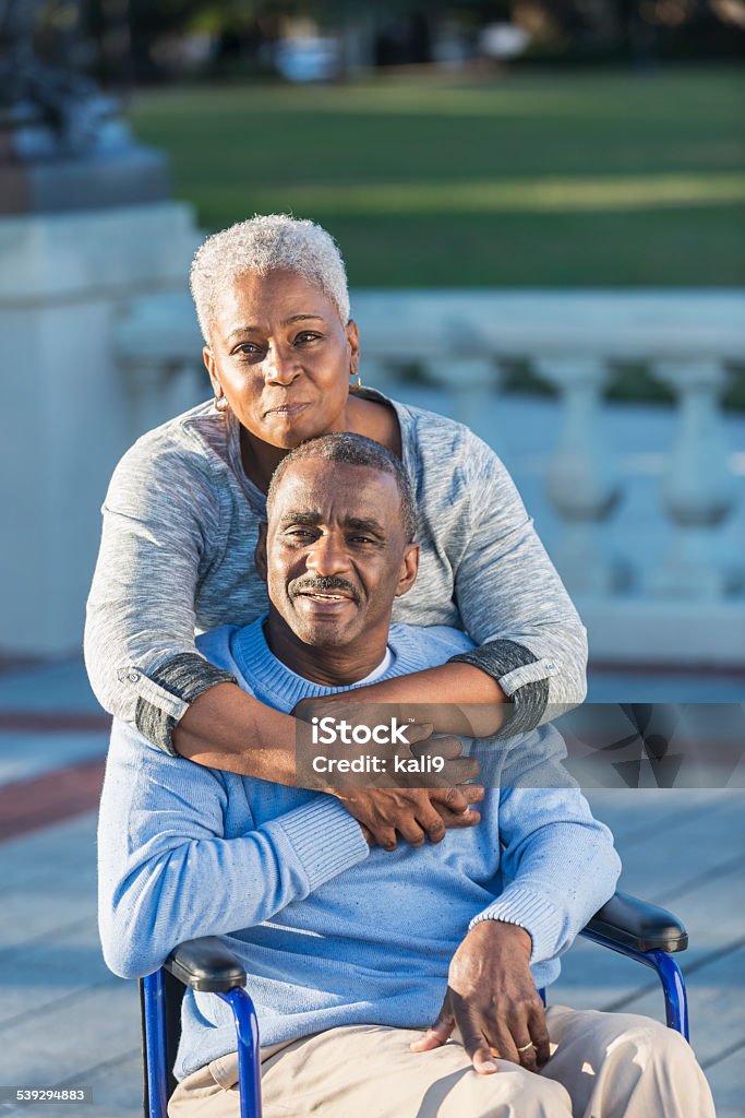 Loving senior couple with man in wheelchair An affectionate senior African American couple at the park. The man is sitting in a wheelchair.  His wife is standing behind him, embracing him, looking at the camera Senior Couple Stock Photo