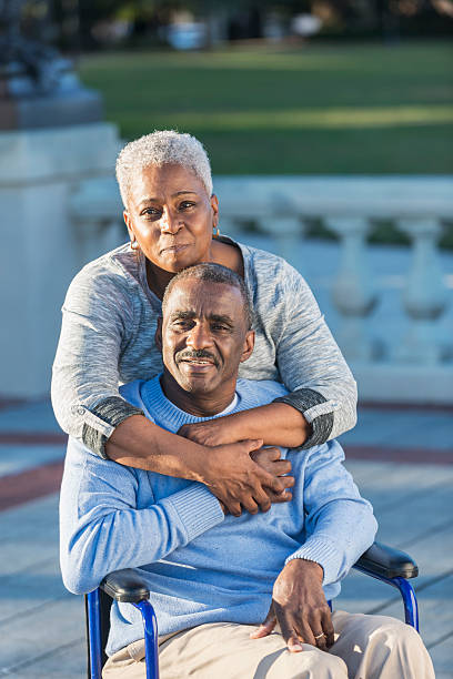 Loving senior couple with man in wheelchair An affectionate senior African American couple at the park. The man is sitting in a wheelchair.  His wife is standing behind him, embracing him, looking at the camera 60 69 years stock pictures, royalty-free photos & images