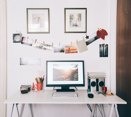 designer's workspace, computer on a white table, photographs hanging on the wall, vintage fashioned, framed paintings on the wall, editing of a photograph in progress on the screen.