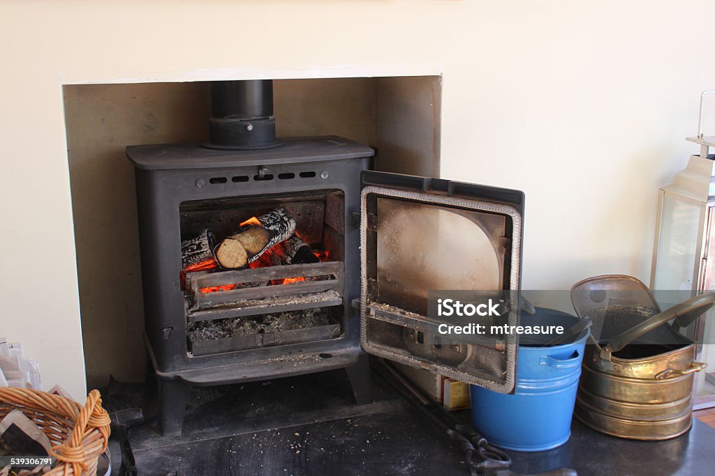 Image of modern fireplace, woodburning stove, coal buckets, log basket Photo showing a traditional cast iron woodburner, pictured with its door open to reveal the logs and fire burning inside.  The metal wood burning stove provides a focal point for a sitting room / lounge, with the modern fireplace simply consisting of a hole in the wall, without an actual mantelpiece surround. Fireplace Stock Photo