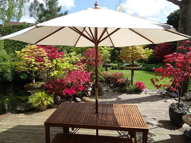 Photo showing a landscaped back garden, which is centred around a spacious area of timber decking, where a wooden table and bench seating is shaded by a cream parasol umbrella.