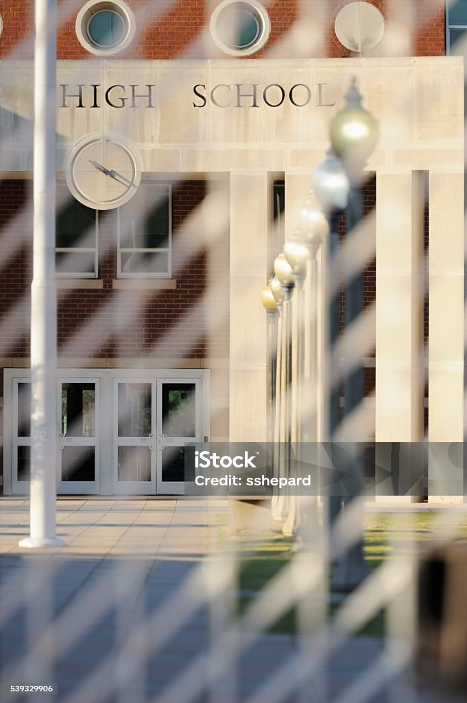 High school with heavy duty security gate fence Looking through heavy duty security gate or fence at entrance way to high school with sign.  Photograph taken thru gate with blurry gate and row of lights leading to entrance. Education Stock Photo