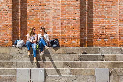 Venice, Italy - June 3, 2016: Two girl friends sit and chat on the steps of the bell tower on a sunny in St Mark's Square, Venice, Italy.